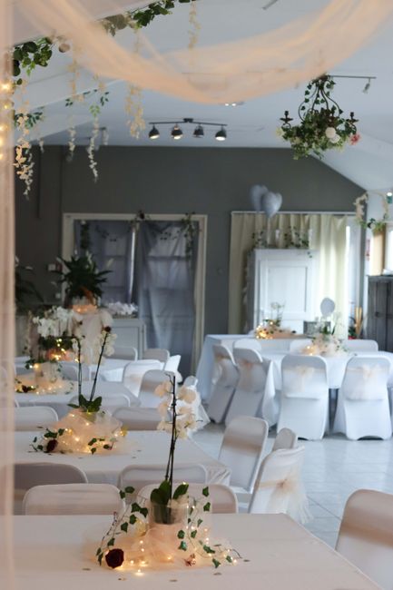 A floral arrangement featuring tall white orchids and dark burgundy roses in a glass vase, placed on a table adorned with a white tablecloth, delicate fairy lights, and scattered flowers. The setting 