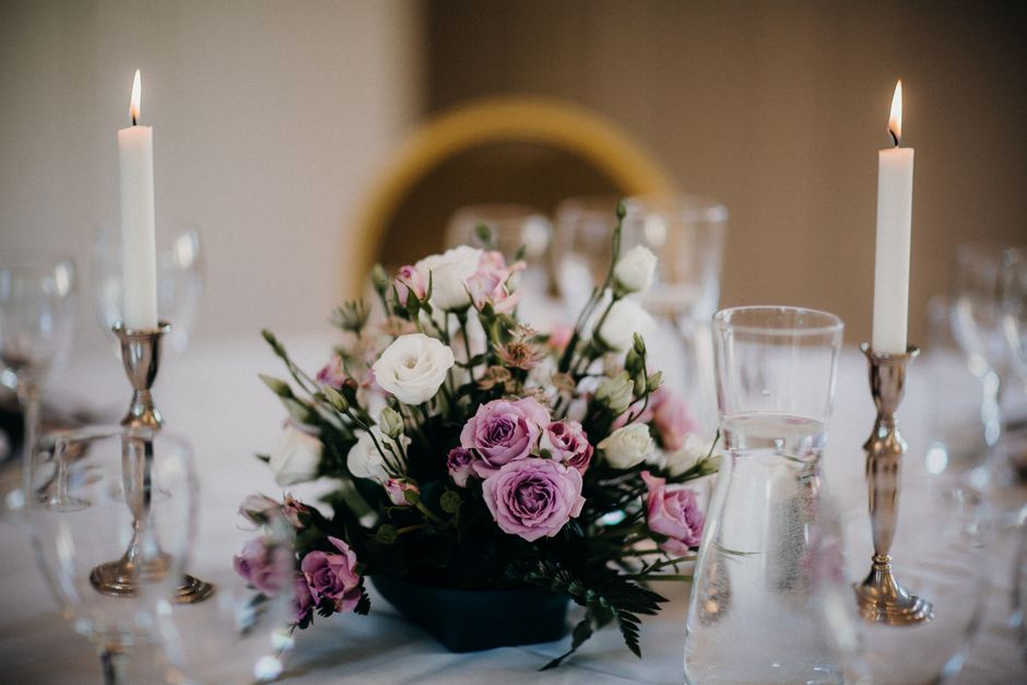 A floral arrangement featuring pink roses and white lisianthus, arranged in a black vase, placed as a centerpiece on a dining table. The bouquet includes lush green foliage and is positioned between t