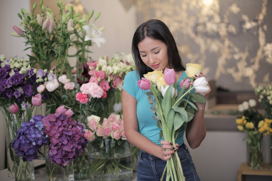 A young woman with shoulder-length dark hair wearing a light blue t-shirt is holding a bouquet of fresh tulips, including pink, white, and yellow blooms, in a florist shop. She is standing amid an ass