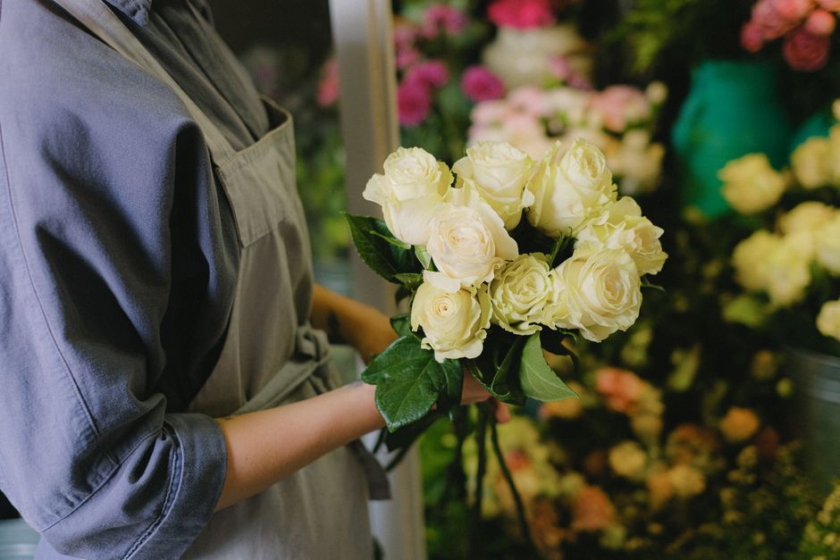 A person wearing a gray apron holds a bouquet of fresh, creamy white roses with lush green leaves, positioned in front of a floral display with various colorful flowers in the background at a flower s