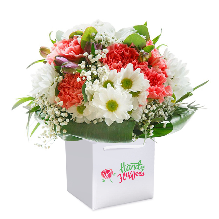 A perfectly balanced bouquet, lovingly composed in Yeading, rises gracefully from a modern white gift bag, its composition as inviting as an open meadow in early summer. Lush carnations shine in shades of coral, peach, and vivid red, their ruffled petals adding texture and layers, while crisp white daisies with lime-green centers lend brightness and cheer. Baby's breath, with its airy clusters of miniature white blooms, fills the spaces between, giving a sense of lightness and movement. Unexpected glimpses of closed buds with subtle strokes of purple on the left side introduce a hint of mystery and anticipation, promising continued vitality. The entire bouquet is encircled by deep, glossy green leaves that act as a natural frame, seamlessly integrating all elements into a cohesive and abundant shape. Sturdy white ribbon handles peek from the gift bag's top, hinting at easy, celebratory delivery through Yeading's neighbourhoods-from Northolt to the peaceful paths of Minet Park. Set against a flawless white background, every detail is bathed in even light, ensuring the freshness and beauty of the bouquet are at the forefront. Designed for thoughtful gifting-be it a birthday surprise, anniversary delight, or a spontaneous gesture-this arrangement reflects the dedication and professionalism of Yeading's local florists.