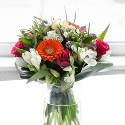 This vivid bouquet, crafted by Yeading florists familiar with the rhythms of local life, sings with exuberant color and texture. Creamy peach and lively orange gerberas form the core, their elegant petals radiating outwards, reminiscent of early morning sunlight on Yeading's residential avenues. Against this cheerful foundation, magenta roses add luxurious depth, their whorled petals a visual embrace of romance and sophistication. Sprinkled throughout are pristine white alstroemeria and delicate spray roses, offering balance and a touch of subtle fragrance, much like white-washed blossoms lining Central Avenue. Light pink accents and tiny white filler flowers peek between verdant layers, giving the arrangement a layered, abundant sense-lush but never overwhelming. The florists' expertise shows in the way broad, glossy aspidistra leaves are artfully wrapped into a verdant collar, providing form and focus. Smaller, pointed leaves and scrolls of greenery add lively contrast, binding the ensemble together. All the elements are anchored in a tall, clear glass vase, the stems perfectly arranged and visible, lending a sense of freshness and authenticity. Soft, ambient lighting brings the colours alive against a minimalist background-every petal, leaf, and nuance clearly seen. Suitable for celebrations, milestones, or sending comfort, this arrangement epitomizes Yeading's balance of vibrancy and tranquility.